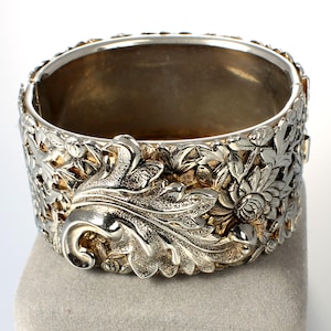 May include: A silver-toned bangle bracelet with an intricate floral design. The bracelet is made of metal and has a hinged clasp.