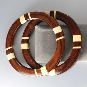 May include: Two wooden bangles with a dark brown finish and cream-colored rectangular inlays. The bangles are circular and appear to be made of a polished wood. The design is simple and elegant, suitable for everyday wear.