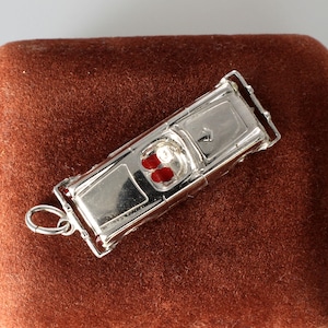 May include: A silver-colored car charm with red accents. The charm is a miniature replica of a classic car, featuring detailed windows and a loop for attachment. The car's interior has two red dots. The charm is on a brown surface.