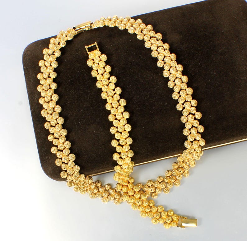K&ouml;nnte beinhalten: Ein goldfarbenes Halsketten- und Armband-Set mit einem zarten, strukturierten Kugelketten-Design.