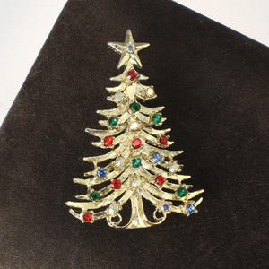 May include: Gold-toned Christmas tree brooch with a star on top. The tree is adorned with colorful rhinestones in red, green, blue, and clear. A festive accessory for the holiday season.