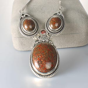 May include: A silver-toned necklace featuring a large, oval pendant with a brown and red patterned stone. Two smaller, matching stones are set above the pendant, with a small red stone accent. The necklace has a delicate chain and ornate metalwork.