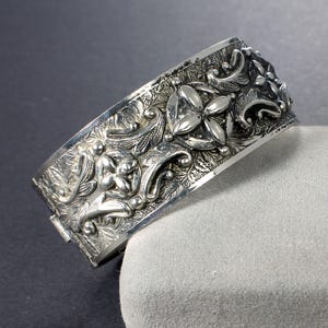 May include: A silver bangle bracelet with an intricate floral design. The bracelet is made of a thick, solid metal and has a hinged clasp.