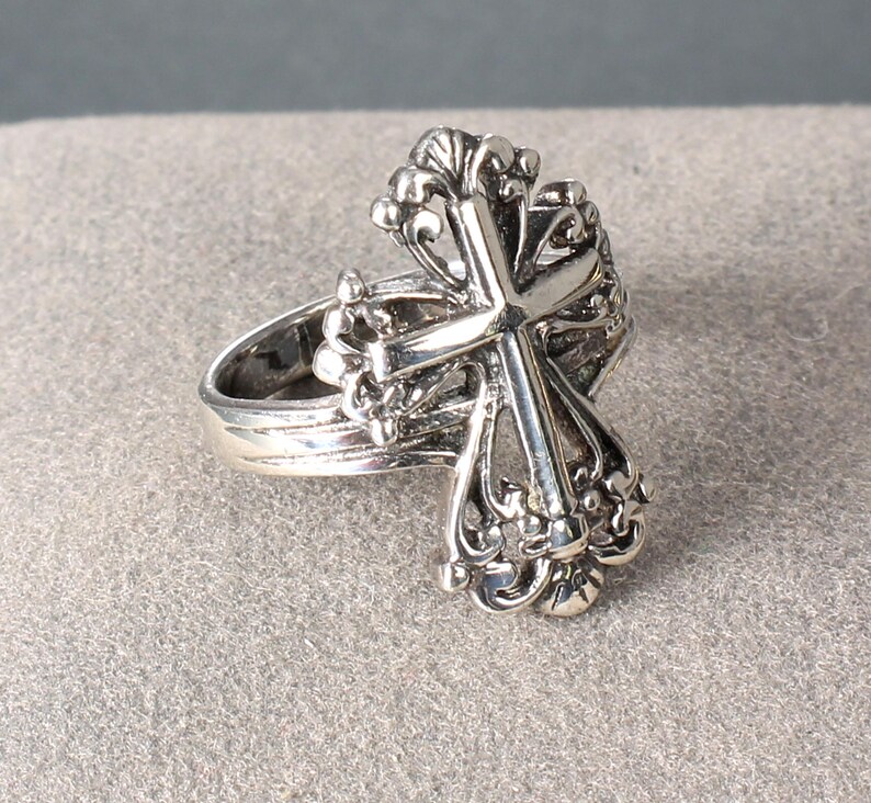 Peut inclure: Une bague argent&eacute;e orn&eacute;e d'une croix d&eacute;taill&eacute;e. La croix est orn&eacute;e de volutes et d'ornements complexes. Le corps de la bague est lisse, contrastant avec la croix &eacute;labor&eacute;e. La bague est probablement en argent sterling. Un bijou d&eacute;coratif.