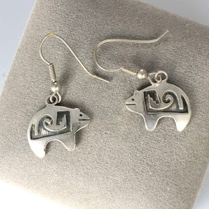 May include: A pair of silver-toned bear-shaped dangle earrings. Each earring features a bear silhouette with an etched design and a hook for pierced ears. The earrings are displayed on a gray surface.