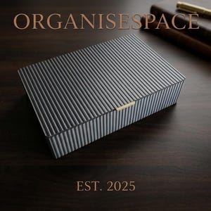Peut inclure: Une boîte de rangement rectangulaire avec un motif à rayures verticales gris foncé et blanc, dotée d'un petit fermoir en métal doré sur le bord avant. Le texte ORGANISESPACE et EST. 2025 apparaît avec un stylo doré et un carnet.