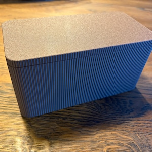 Könnte beinhalten: Eine rechteckige Box mit einem strukturierten, hellbraunen Deckel und einem blauen, vertikal gerippten Körper. Die Box steht auf einer Holzoberfläche, der Deckel ist etwas dunkler als der Körper.
