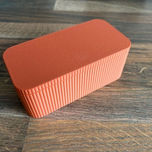 Könnte beinhalten: Eine orangefarbene rechteckige Box mit strukturiertem Deckel und geripptem, vertikalem Design an den Seiten. Die Box besteht aus einem festen Material und steht auf einer Holzoberfläche.