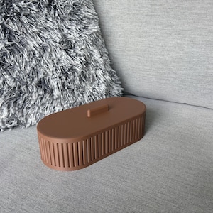 Puede incluir: Una caja marrón de forma ovalada con tapa y laterales acanalados verticales. La caja está hecha de un material sólido y se encuentra sobre una superficie gris. Una almohada gris y esponjosa está en el fondo.