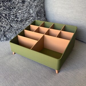 May include: A square olive green desk organiser featuring a ribbed exterior texture and four small light-coloured cylindrical feet. The interior is divided into nine compartments of varying sizes by light-coloured vertical dividers.