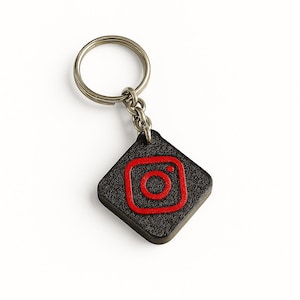 以下が含まれることがあります： 黒と赤のInstagramロゴキーホルダー。キーホルダーは、テクスチャ加工された表面を持つ正方形のチャームが特徴です。Instagramのロゴは赤色で、キーホルダーにはシルバーの金属リングが付いています。
