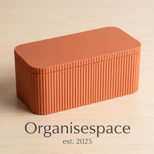 Op de afbeelding: Een rechthoekige oranje opbergdoos met een getextureerd deksel en verticaal geribbelde zijkanten. De doos staat op een licht houten oppervlak. De tekst "Organisespace est. 2025" staat onderaan.