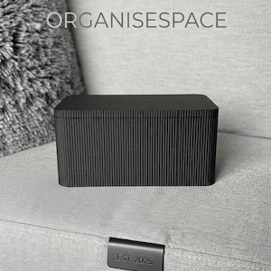 Könnte beinhalten: Eine schwarze, rechteckige Aufbewahrungsbox mit vertikalen Rillen. Die Box steht auf einer grauen Oberfläche. Der Text "ORGANISESPACE" befindet sich über der Box. Der Text "EST. 2025" steht auf einem kleinen schwarzen Etikett.