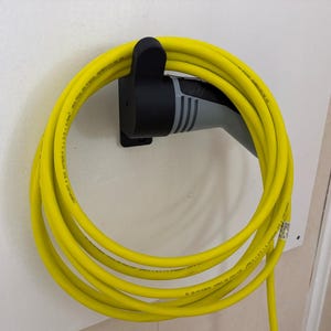 Könnte beinhalten: Ein gelbes Ladekabel für Elektrofahrzeuge, auf einer schwarzen Wandhalterung aufgerollt. Der Stecker ist grau und schwarz. Die Wand ist weiß.