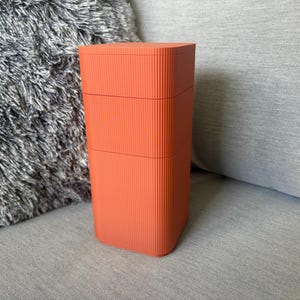 Puede incluir: Un recipiente de almacenamiento rectangular y alto de color naranja con una textura vertical acanalada en todos sus lados. El objeto cuenta con una tapa plana y dos costuras horizontales, lo que indica un diseño modular o apilable.