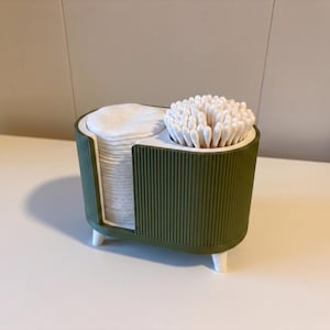 Op de afbeelding: Een olijfgroene badkamerorganizer met een geribbelde buitenkant en witte pootjes. De houder heeft twee vakken: één bevat een stapel ronde witte wattenschijfjes en de andere bevat een bundel witte wattenstaafjes voor persoonlijke verzorging.