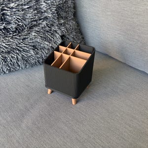 Pode incluir: Um organizador de secretária retangular com um exterior preto canelado e um interior castanho claro com vários compartimentos. O organizador é suportado por quatro pequenas pernas cónicas castanho claro que elevam a base.