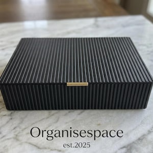 May include: A black rectangular storage box with a ribbed texture and a gold-colored handle. The box is sitting on a marble surface. The text "Organisespace est.2025" is at the bottom.
