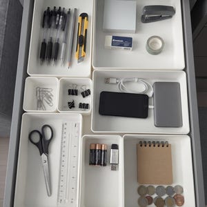 Pode incluir: Uma gaveta de secretária cinzenta organizada com tabuleiros de plástico branco contendo material de escritório, incluindo canetas pretas, um x-ato amarelo, um agrafador, clipes, molas, tesouras, uma régua de 15 cm, pilhas, um smartphone e moedas.