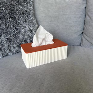 Puede incluir: Un dispensador de pañuelos rectangular, blanco con ranuras verticales y tapa marrón rojizo. Los pañuelos blancos sobresalen de la abertura. El dispensador está sobre una superficie gris, cerca de un cojín gris.