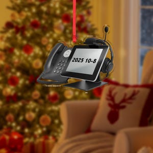 May include: A Christmas ornament featuring a black telephone, a tablet with the text "2025 10-8", and a headset. The ornament hangs from a red ribbon, with a blurred Christmas tree in the background. The ornament is a unique holiday decoration.