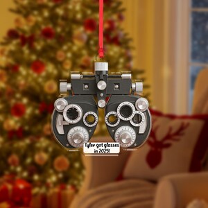 以下が含まれることがあります： 赤いリボンが付いた、黒とシルバーのフォロプターのクリスマスオーナメント。オーナメントには複数のダイヤルとレンズがあります。小さな白い看板には「Tyler got glasses in 2025！」と書かれています。