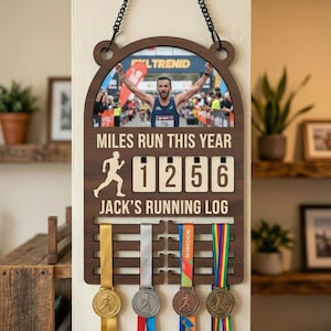 以下が含まれることがあります： ランナーの写真と「MILES RUN THIS YEAR」と「1256」マイルを表示する木製のランニングログ。メダル用のスペースと「JACK'S RUNNING LOG」のテキストが含まれています。ログは黒いチェーンから吊り下げられています。