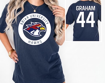 Könnte beinhalten: Marineblaues T-Shirt mit dem Logo der Briar University Hawks auf der Vorderseite. Das Logo ist ein weißer Kreis mit den Worten "Briar University" und "Hawks" um einen stilisierten Falken. Auf der Rückseite des Shirts stehen "GRAHAM" und die Nummer "44" in Weiß.