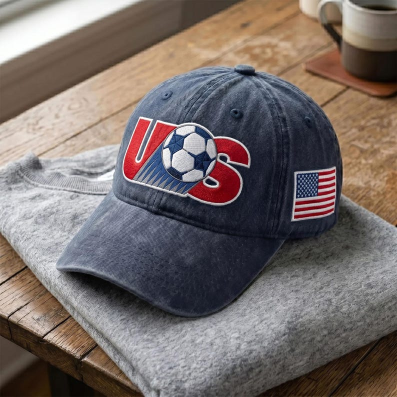 May include: A navy blue baseball cap featuring an embroidered graphic on the front with the letters US in red and a soccer ball replacing the letter S. A small rectangular patch of the United States flag is stitched onto the side of the cap.