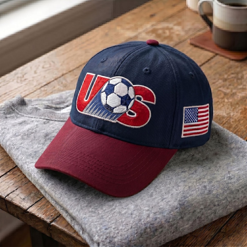 May include: A navy blue baseball cap with a maroon brim. The front features an embroidered red 'US' logo with a soccer ball replacing the letter 'S'. A small American flag patch is embroidered on the side of the cap, which has a button on top.