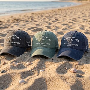 May include: Three baseball caps in dark grey, green, and navy blue, each with the text "FATHERHOOD IS KINGDOM WORK Proverbs 22:6" and a cross graphic. The caps are on a sandy beach.
