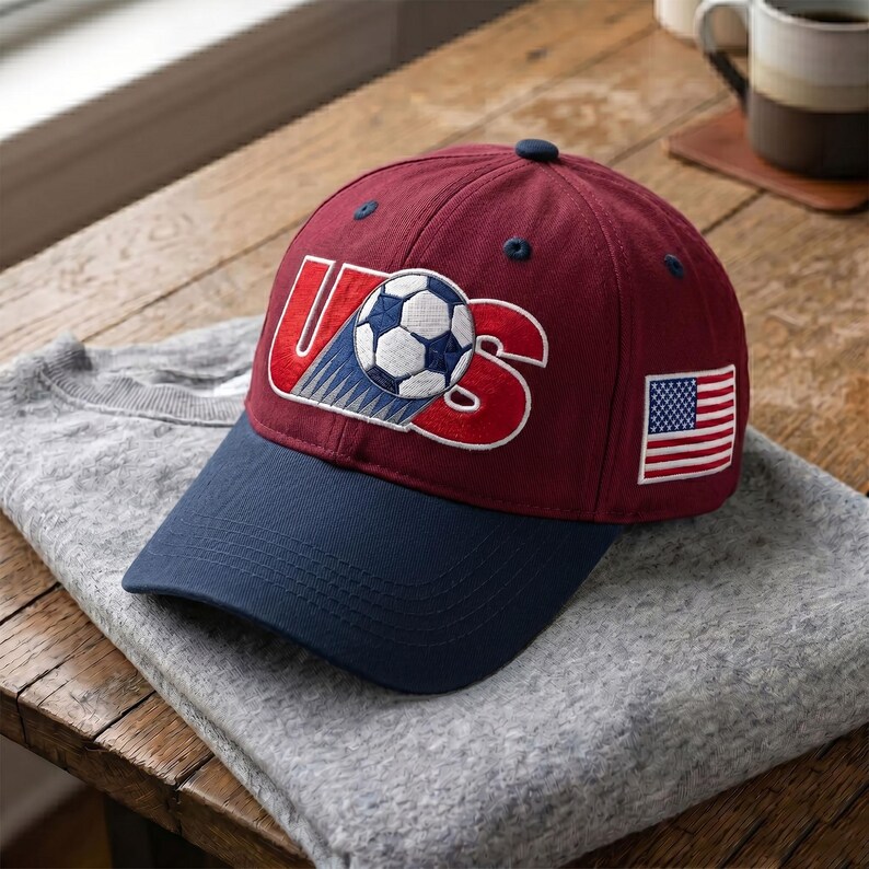 May include: A maroon baseball cap with a navy blue brim. The front features an embroidered graphic with the letters US in red and a soccer ball in the center. A small embroidered American flag is on the side of the cap, which has navy blue eyelets.