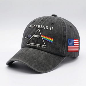 May include: A dark gray baseball cap with the text "ARTEMIS II" and "DARK SIDE OF THE MOON". The cap features a triangle design with a rainbow and a small American flag patch. The cap is made of a worn, textured material.