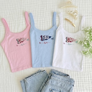 May include: Three tank tops in pink, light blue, and white, each with a graphic design. The pink and white tops feature "Stanford" text, while the blue top says "FAD". All have a bow and the name "Taylor" below. Light wash denim jeans are at the bottom.