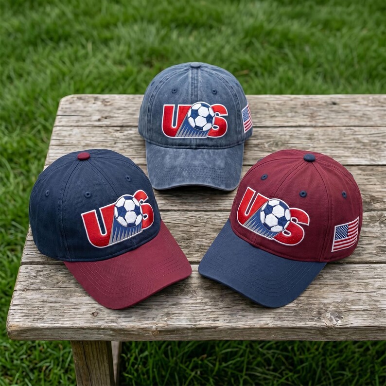 May include: Three baseball caps featuring embroidered soccer ball graphics and the text US. One cap is blue with a red brim, one is maroon with a blue brim, and one is a faded blue denim style. Each cap displays a small American flag on the side.