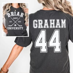 May include: Dark gray t-shirt with white text and graphics. The front features "BRIAR HAWKS UNIVERSITY" with crossed hockey sticks and a puck. The back displays "GRAHAM 44" in a collegiate font. The shirt is a comfortable, casual style.