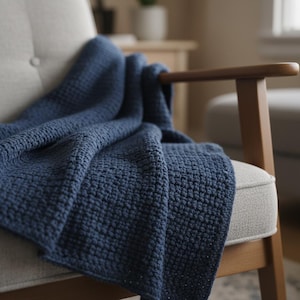 May include: A navy blue knitted throw blanket draped over a light gray armchair. The blanket has a textured, woven pattern. The chair has wooden armrests and a light gray seat and back. The background is softly blurred.
