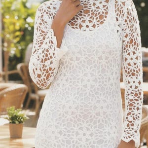 Könnte beinhalten: Weißes, knielanges Häkelkleid mit langen Ärmeln. Das Kleid hat ein Blumenmuster und einen runden Ausschnitt. Das Model trägt das Kleid im Freien, mit einem verschwommenen Hintergrund aus Grün und Tischen.