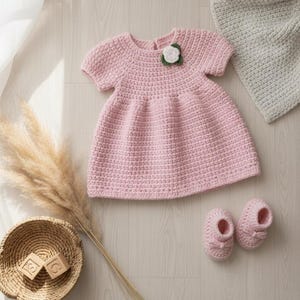 以下が含まれることがあります： 手作りのピンク色のベビー用かぎ針編みドレス。短い袖とフレアスカートがあり、白い花のアップリケで飾られています。お揃いのピンクのかぎ針編みベビーブーティーが近くにあります。薄い灰色の毛布と木製のブロックが入った籐のバスケットも画像にあります。