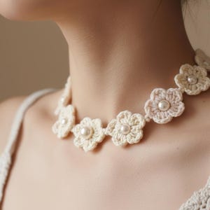 Puede incluir: Un collar de flores de ganchillo color crema con detalles de perlas. El collar presenta múltiples formas de flores de ganchillo, cada una con una perla central. El collar se lleva alrededor del cuello.