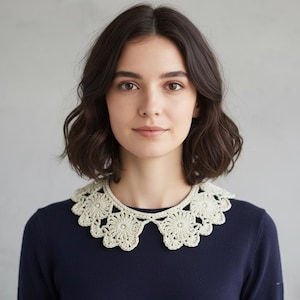 May include: A cream-colored, crocheted collar with a floral design and small pearl accents. The collar is worn over a navy blue sweater. The collar is a handmade accessory.