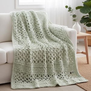 May include: A light green crochet blanket draped over a white sofa. The blanket features a textured, open weave design. The soft, pastel color and open weave create a cozy and inviting aesthetic. The blanket is a home decor item.