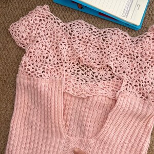 May include: A light pink, sleeveless top with a crochet yoke. The yoke features a floral pattern, and the body of the top is a ribbed knit. The top is made of a soft, lightweight material, perfect for warm weather.