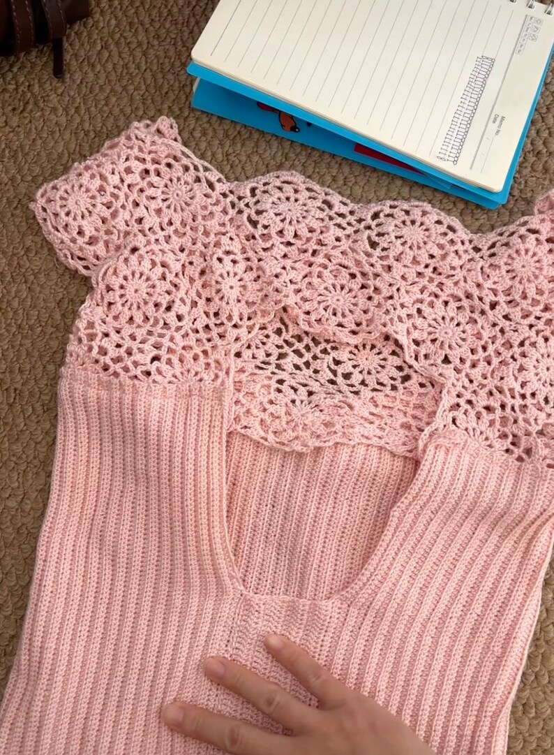 May include: A light pink, sleeveless top with a crochet yoke. The yoke features a floral pattern, and the body of the top is a ribbed knit. The top is made of a soft, lightweight material, perfect for warm weather.