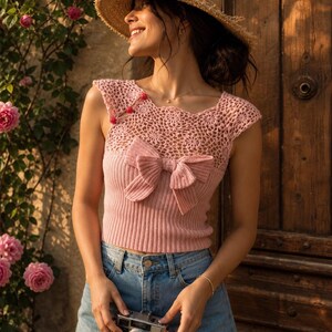 May include: A light pink knitted top with a bow detail and a straw hat. The top features a crochet neckline and is paired with denim shorts. A vintage camera is held in the hands.