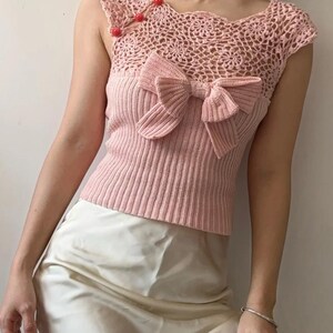 May include: A light pink knitted top with a large bow detail on the front. The top features a crochet neckline and shoulder straps with three red button closures. The bottom portion of the top is ribbed.