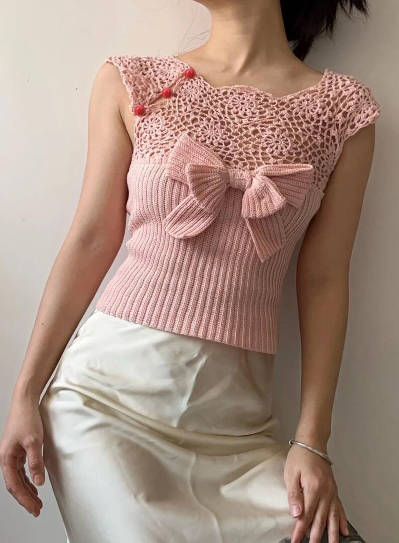 May include: A light pink knitted top with a large bow detail on the front. The top features a crochet neckline and shoulder straps with three red button closures. The bottom portion of the top is ribbed.