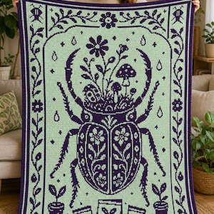 Op de afbeelding: Een rechthoekige gehaakte deken met een licht mintgroene achtergrond, een donkerpaarse rand en een centraal ontwerp. Het patroon toont een grote kever met bloemen en paddenstoelen die uit de rug groeien, omringd door kleine potplanten.