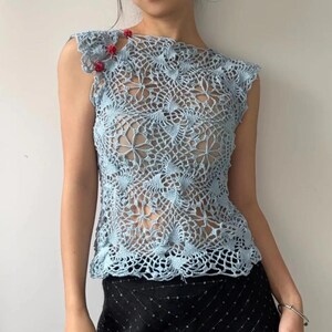 May include: A light blue crochet top with a floral pattern. The sleeveless top features a delicate openwork design and is adorned with small red beads on the shoulder. The top is paired with a black skirt.