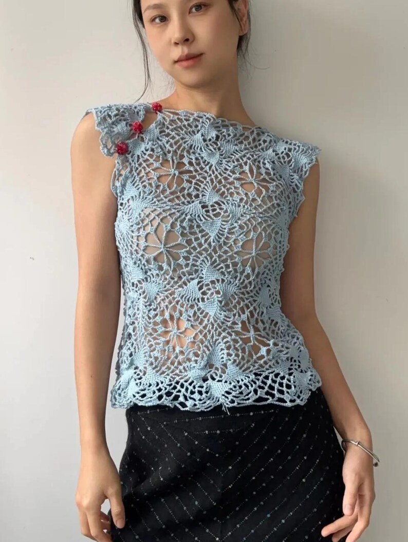 May include: A light blue crochet top with a floral pattern. The sleeveless top features a delicate openwork design and is adorned with small red beads on the shoulder. The top is paired with a black skirt.